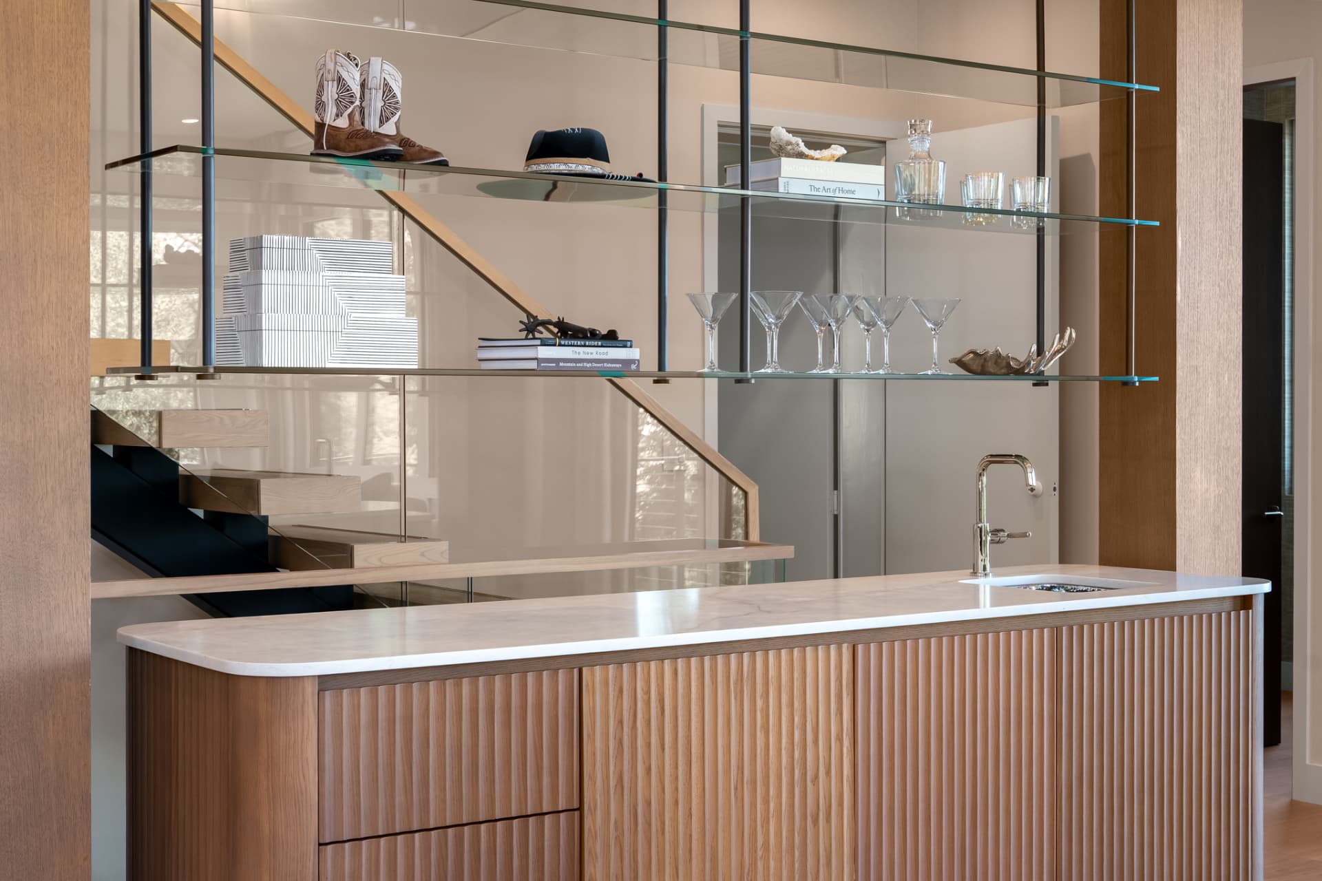 Wet bar with fluted white oak millwork, glass shelving and floating staircase beyond