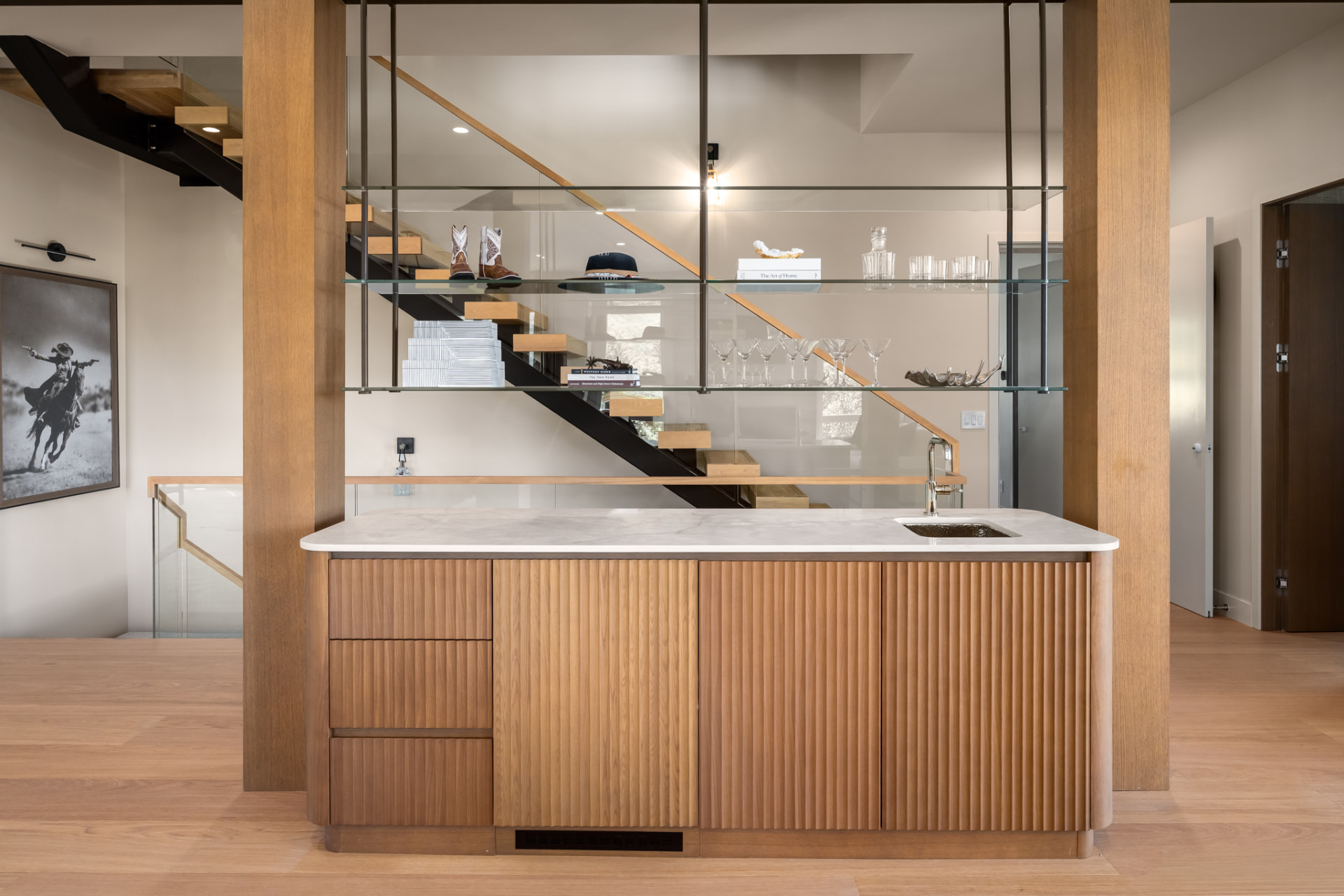 Wet bar with fluted millwork and glass shelving beside the floating staircase